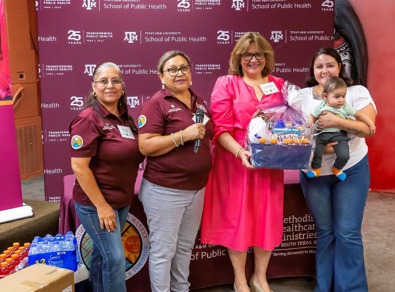 Group of four women and a baby at a public health event, holding a gift basket, in front of a backdrop featuring logos for Texas A&M School of Public Health.
